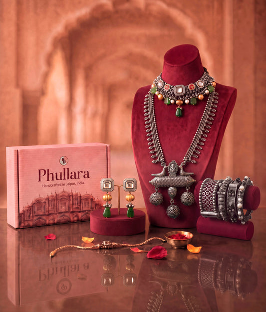 A styled product display of traditional Indian jewelry from Phullara, featuring a silver-toned necklace with green and red stones on a red velvet stand, a matching silver bracelet, earrings, and a gold-toned bracelet, alongside the pink Phullara brand box.